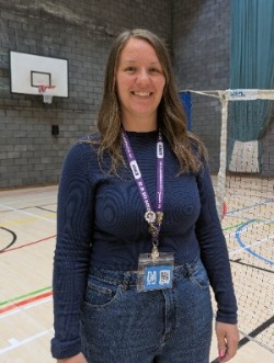 Lauren Simpson smiling and standing in an indoor sports hall in front of a basketball hoop and net.